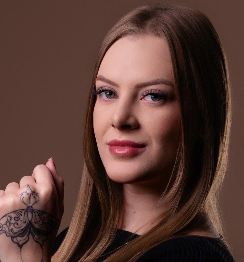 Dra. Camila Domanski contra parede marrom, de blusa preta, com a mão erguida mostrando a tatuagem ornamental