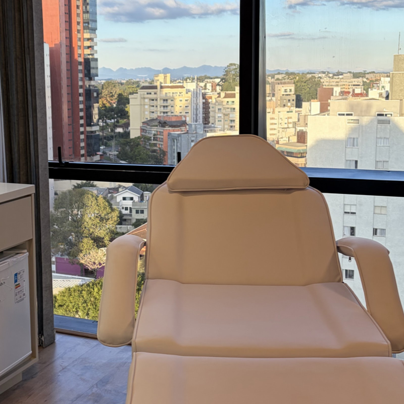 Poltrona de atendimento em tom nude de frente para a janela de esquadria preta, com a cidade de Curitiba e a serra ao fundo