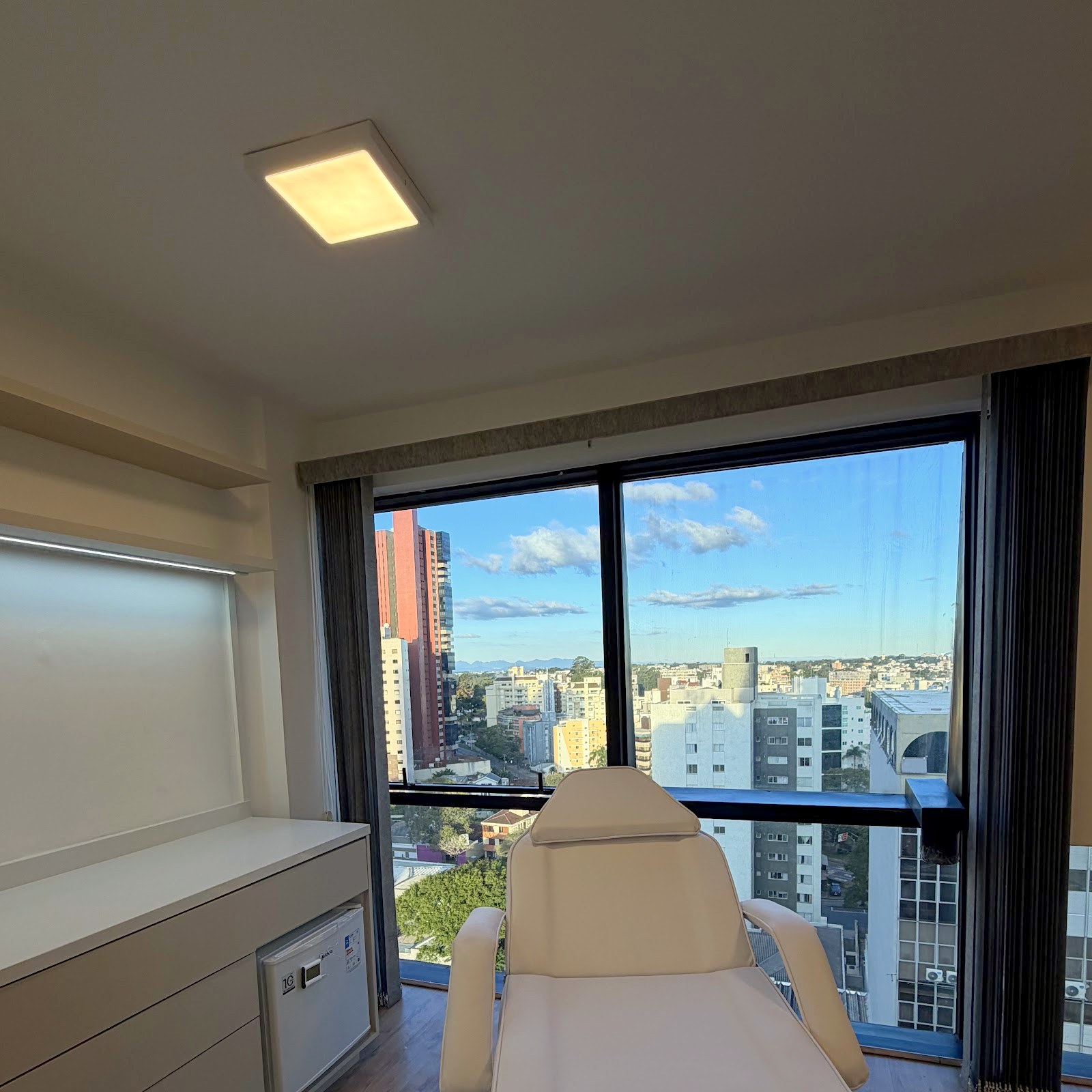 Vista da sala no 12º andar: a janela de esquadria preta emoldurando os prédios de Curitiba, as árvores e o céu com nuvens altas